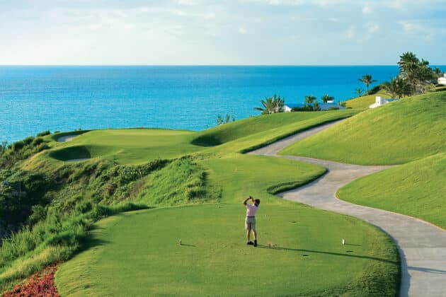 A man is standing on a golf course near the ocean, enjoying his bermuda vacation.