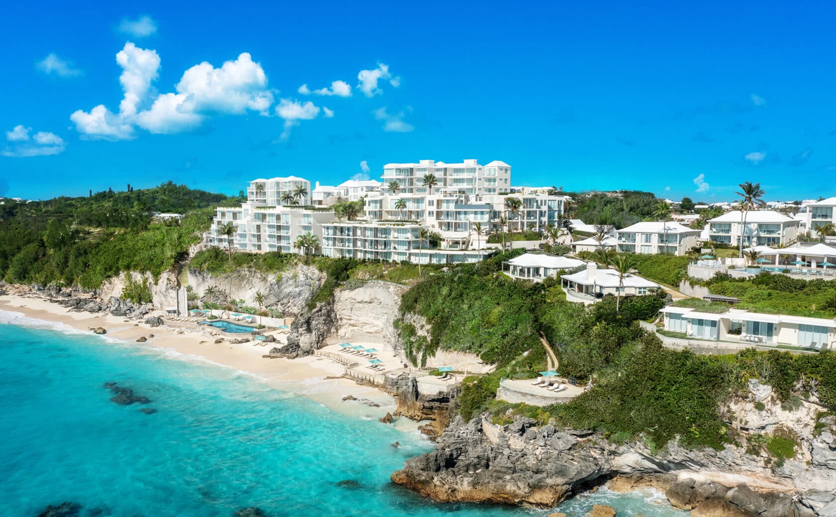Aerial view of a luxurious coastal resort with white buildings perched on rocky cliffs above a turquoise sea, surrounded by lush greenery and a sandy beach.