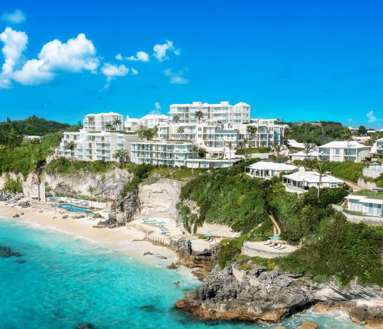 Aerial view of a luxurious coastal resort with white buildings perched on rocky cliffs above a turquoise sea, surrounded by lush greenery and a sandy beach.
