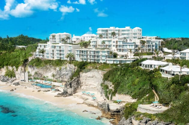 Luxury seaside resort with white buildings nestled on cliff above a turquoise beach with loungers and lush greenery.