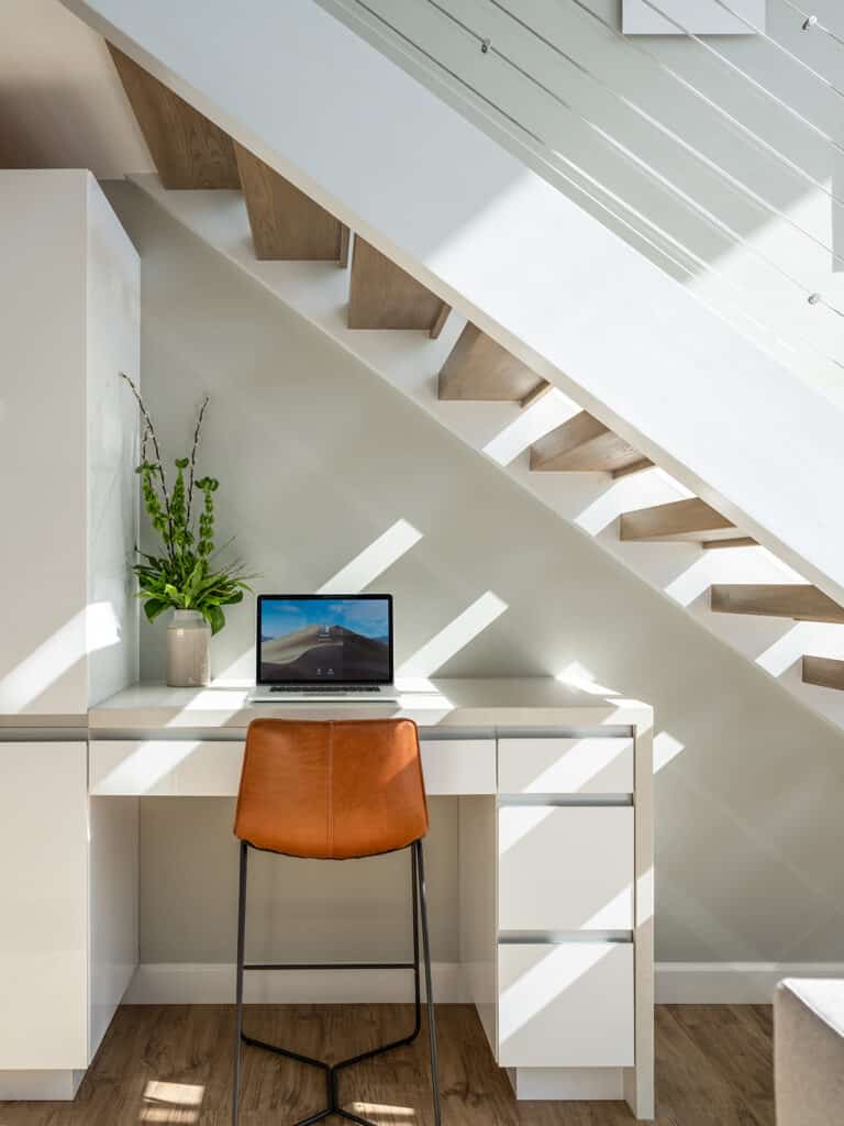 In a modern workspace tucked under a staircase, featuring a desk, laptop, potted plant, and brown chair, sunlight streams through the railing. This stylish setup evokes the charm of Bermuda real estate, where light and design harmonize to enhance any living space.