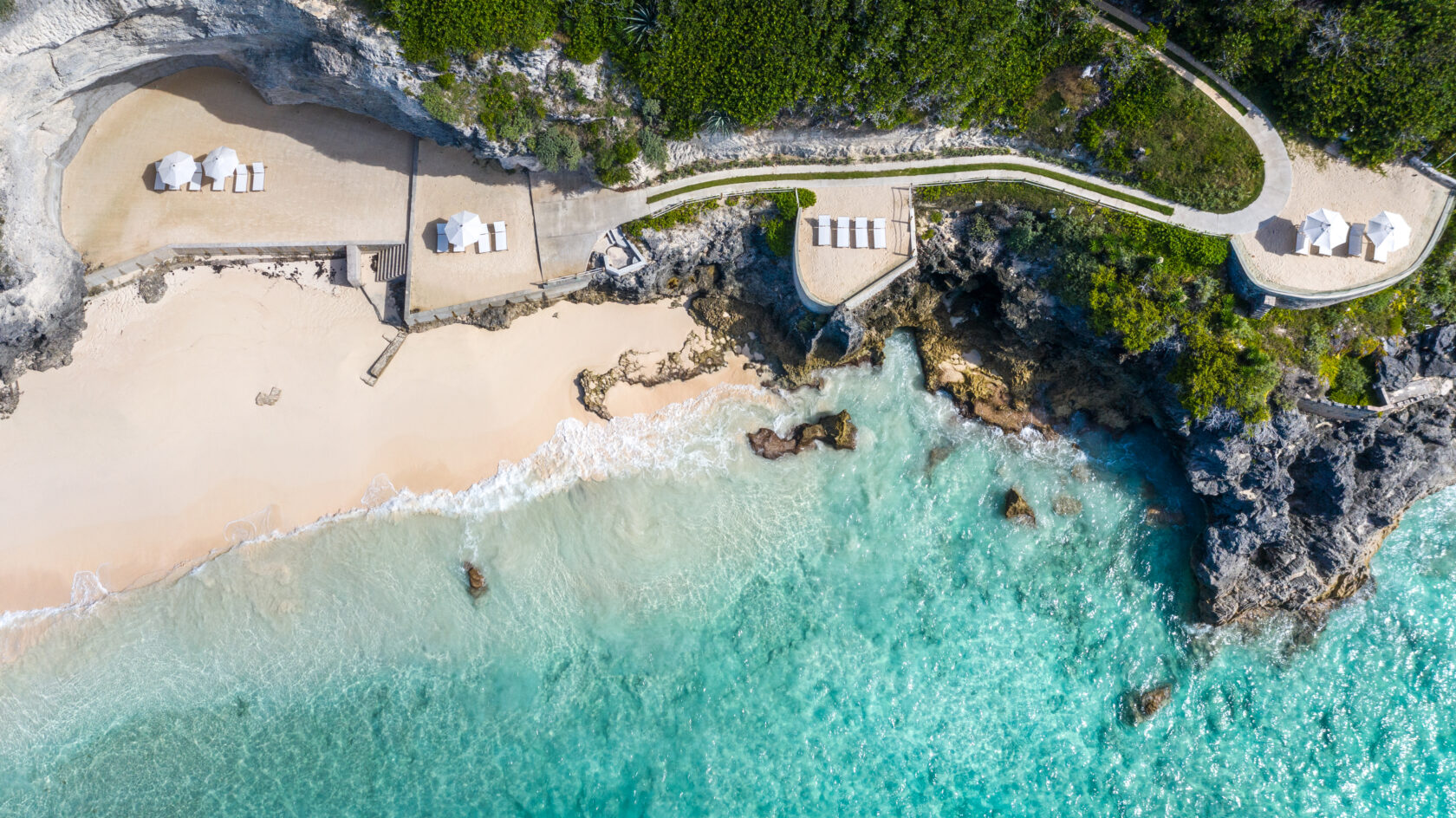 Aerial view of a secluded beach with turquoise water, surrounded by rocky cliffs and greenery. Features include sandy areas, white umbrellas, and a winding path nearby—a perfect spot for those considering Bermuda vacation rentals.