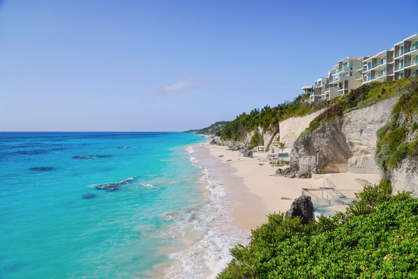 A scenic view of a beach with pristine turquoise water and white sand, bordered by cliffs and modern buildings, under a clear blue sky, showcases the allure of Bermuda vacation rentals nestled along the picturesque coastline.