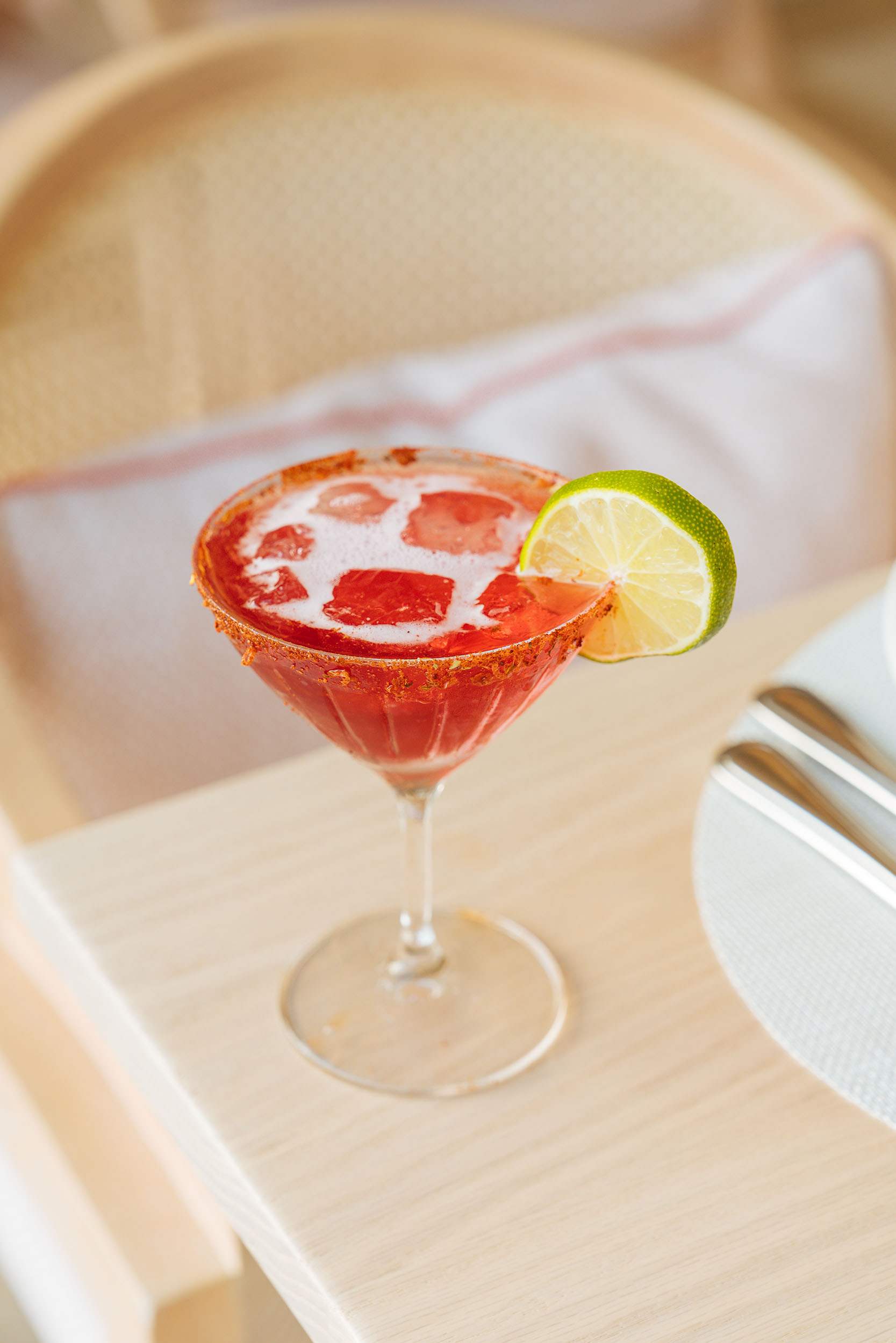 A red cocktail with ice cubes sits in a martini glass, garnished with a lime slice on the rim, evoking the refreshing allure of Bermuda vacation rentals, placed on a light wooden table.