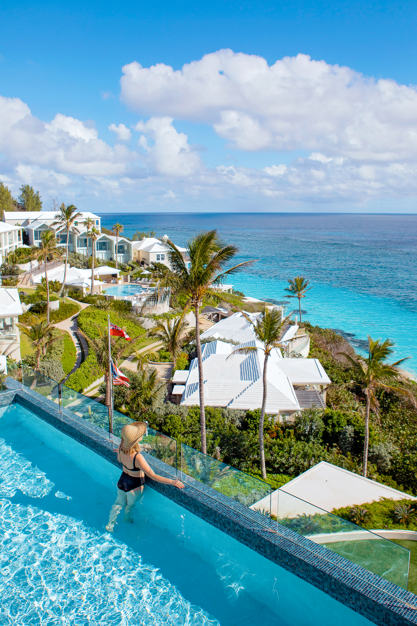 A person in swimwear stands in an infinity pool overlooking a coastal resort with palm trees and ocean views under a cloudy sky, capturing the essence of Bermuda residences and hinting at the allure of long-term stays near Hamilton, Bermuda.