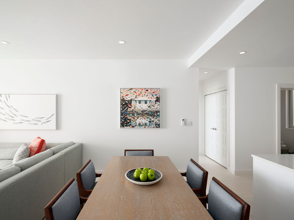 Modern dining area with a wooden table, six chairs, and a bowl of green apples. Mounted artwork on the wall, with a glimpse of a gray sofa on the left. This minimalist and clean design is perfect for long-term stays near Hamilton, Bermuda.