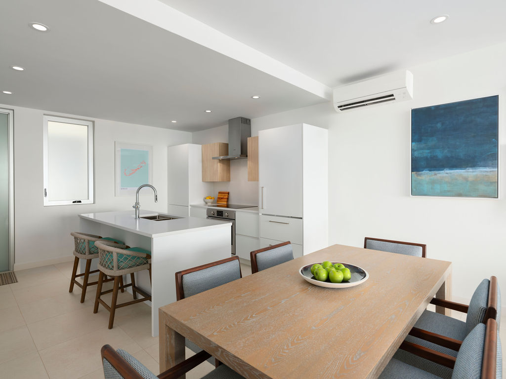 Modern kitchen and dining area in this Bermuda residences listing features white cabinets, a large wooden table, chairs, and a kitchen island with stools. A bowl of green apples sits on the table, complemented by artwork on the wall, exuding an inviting elegance.